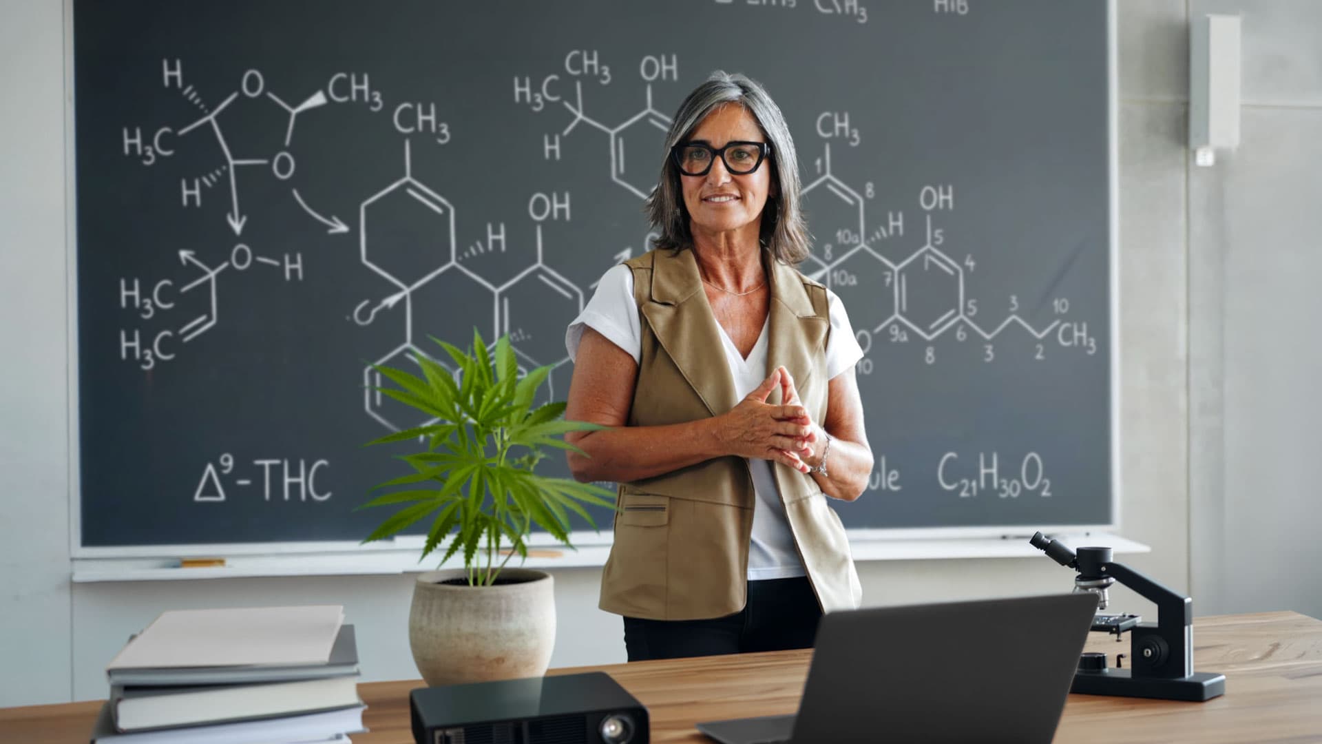 Woman teaching about THC beside a cannabis plant and chemical structures on a chalkboard.