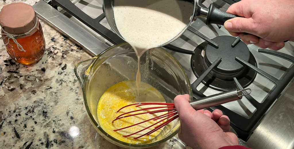 Pouring warm milk mixture into egg yolks while whisking to temper the eggs during eggnog preparation.