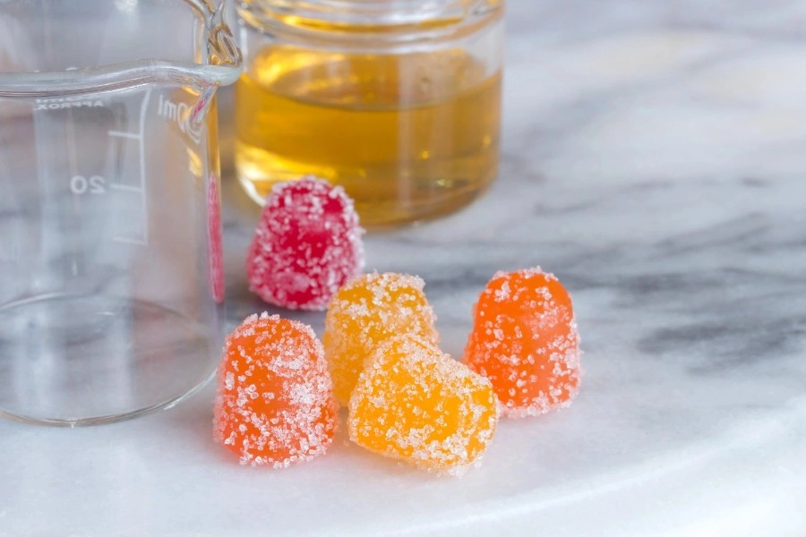 Colorful THC-infused gummies covered in sugar crystals, placed beside a measuring beaker and a small jar of cannabis oil, highlighting the ingredients and dosage process in making potent weed gummies.
