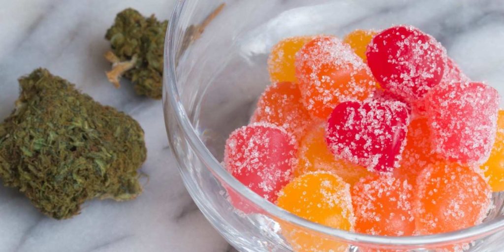 Assorted red, yellow, and orange weed gummies in a glass dish next to dried cannabis buds on a white marble surface