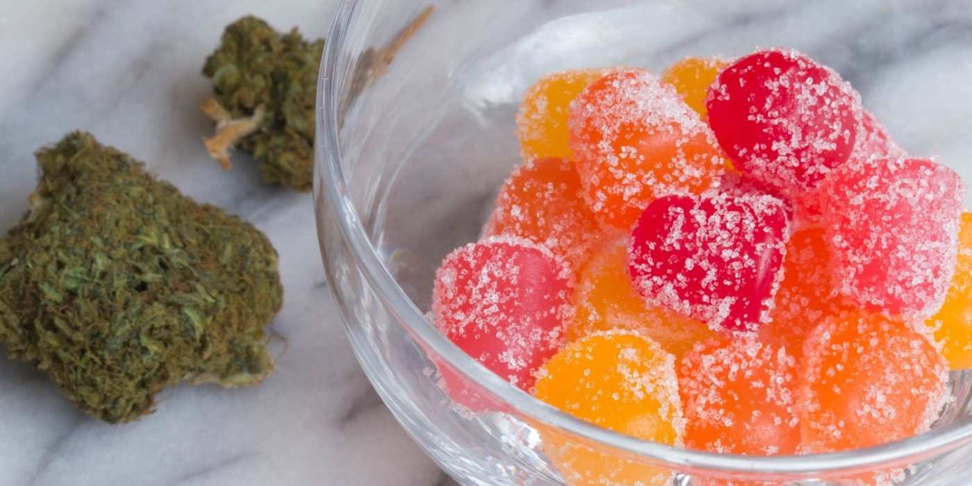 Colorful homemade weed gummies coated in sugar in a glass bowl, with cannabis buds on a marble background