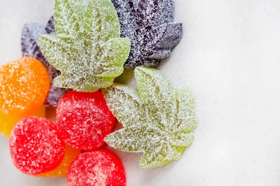 Assorted homemade cannabis gummies, including green leaf-shaped and round red, orange, and purple pieces, all coated in sugar crystals, representing the variety and care in crafting weed edibles.