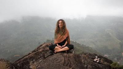 Woman meditating in a seated yoga pose on a rocky overlook surrounded by misty mountains.