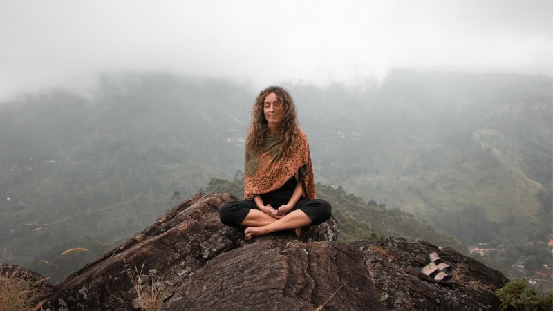 Woman meditating in a seated yoga pose on a rocky overlook surrounded by misty mountains.