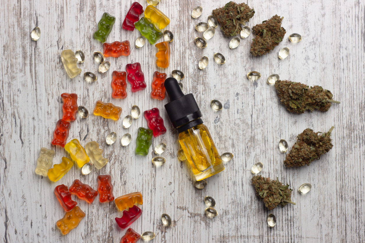 Glass bottle of yellow cannabis CBD oil, dried marijuana buds, cbd capsules, and assorted gummy bears on a rustic wooden table
