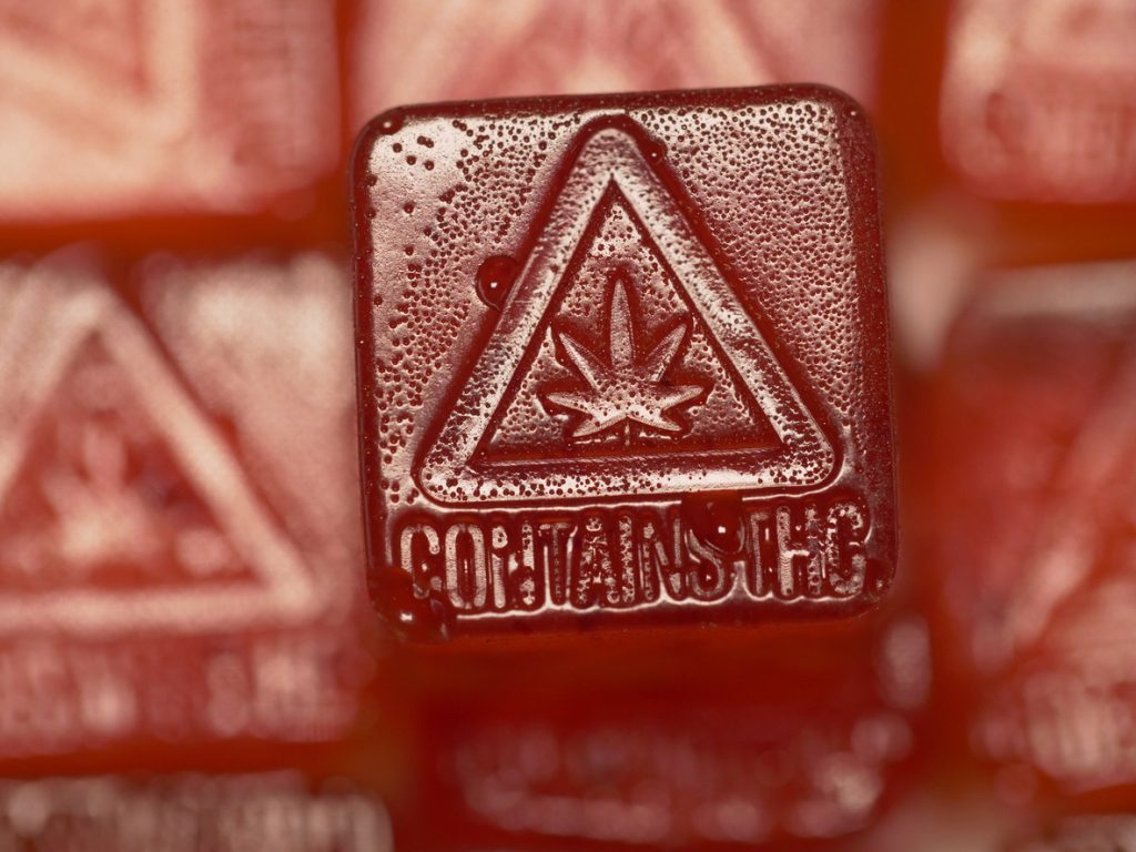 Red cannabis-infused gummy with a triangle warning symbol, cannabis leaf icon, and "Contains THC" text embossed on the surface to indicate it is a THC edible.
