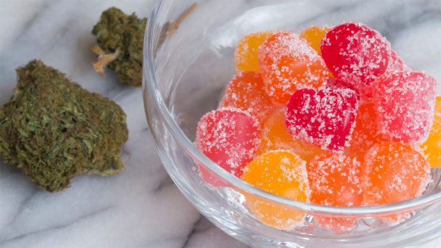 Colorful homemade weed gummies coated in sugar in a glass bowl, with cannabis buds on a marble background