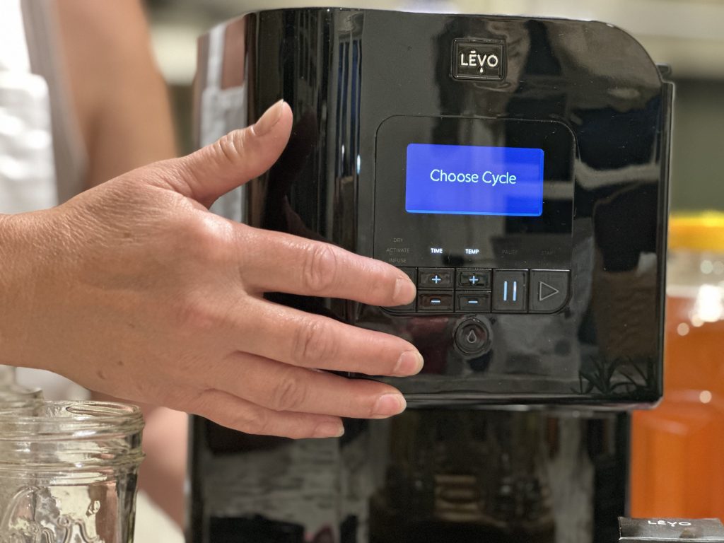 Close-up of the LĒVO infusion machine on a kitchen counter displaying “Choose Cycle,” surrounded by honey, oranges, herbs, and the herb pod, illustrating the setup step for making infused rosemary citrus honey.