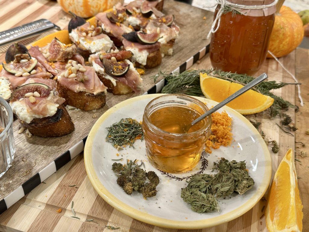 Plate with raw honey, dried rosemary, orange zest, cured cannabis buds, and decarboxylated cannabis displayed beside a jar of honey, with crostini and fresh figs in the background.