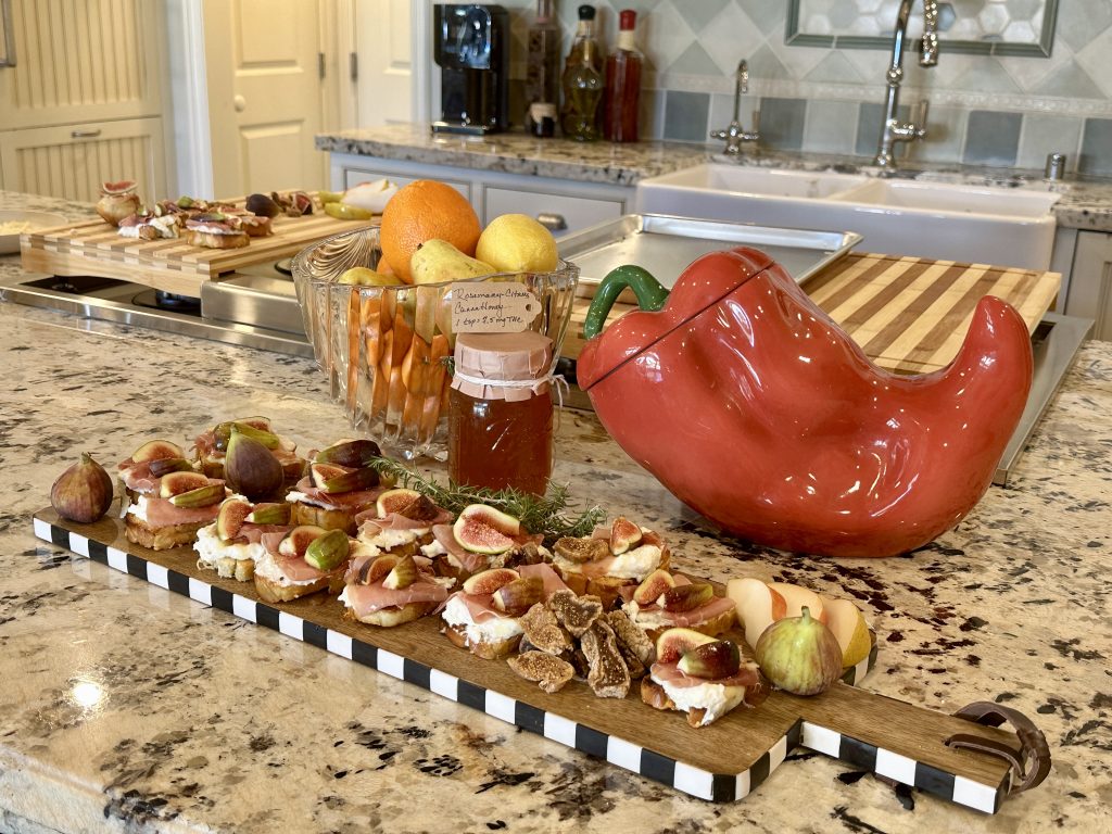 A platter of finished ricotta, prosciutto, and fig crostini drizzled with infused citrus honey, arranged on a kitchen counter with holiday ingredients.