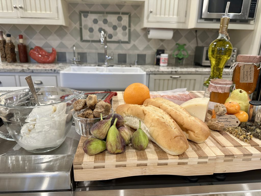 Fresh figs, ricotta, citrus, baguettes, dried figs, infused honey, and olive oil arranged on a wooden board in a home kitchen, ready for assembling ricotta and fig crostini with rosemary, citrus, and cannahoney.