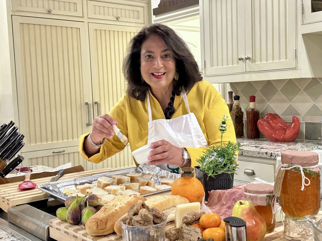 Woman in a yellow top and white apron brushing sliced bread on a baking tray, surrounded by ingredients for infused holiday appetizers including figs, citrus, bread, prosciutto, and jars of homemade cannahoney in a warm kitchen.