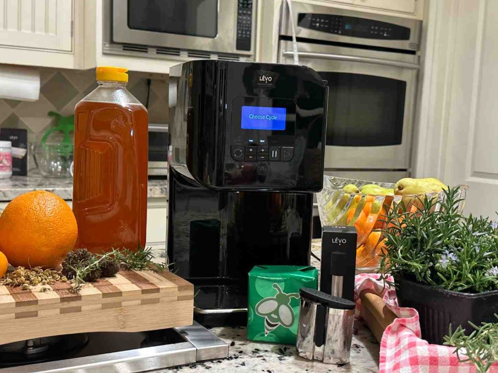 LĒVO infusion machine on a kitchen counter surrounded by fresh rosemary, citrus, honey, and herbs — a clean, precise setup for making homemade cannabis oil, butter, and cannahoney.