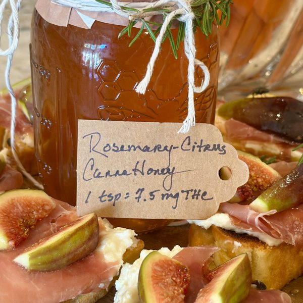 Jar of homemade Rosemary Citrus CannaHoney labeled with dosing information, surrounded by toasted crostini topped with ricotta, prosciutto, fresh figs, and rosemary on a wooden board.