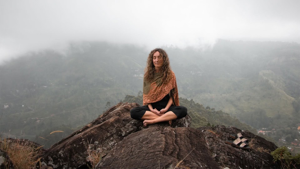 Woman sitting cross-legged in meditation on a rocky hill overlooking misty mountains.