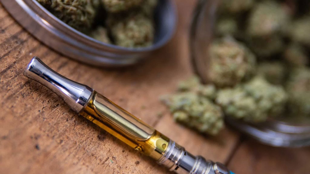 Vape oil cartridge with amber-colored liquid beside cannabis flower on a wooden surface.